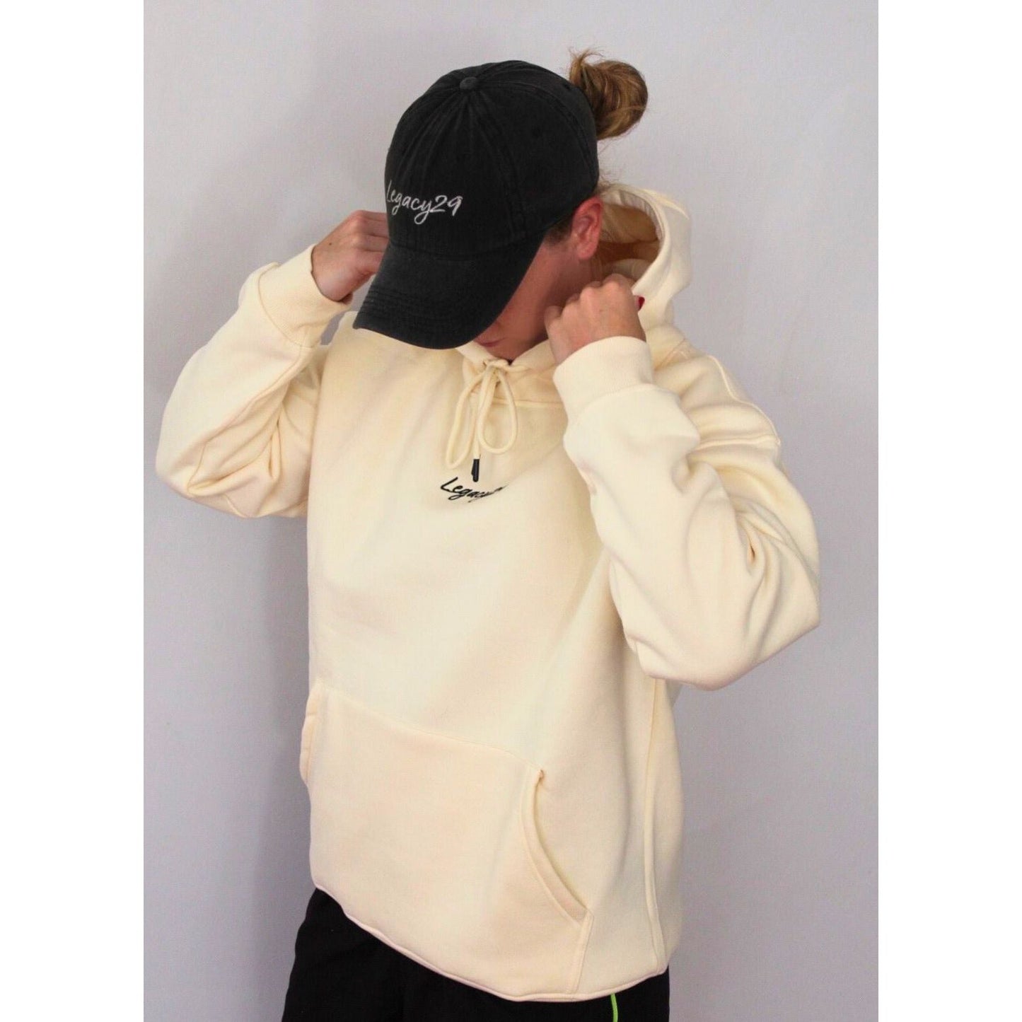 Person wearing Legacy29 Cloud 9 cream hoodie with a black cap, adjusting the hood. Luxurious oversized fit with embroidered logo for a stylish look.
