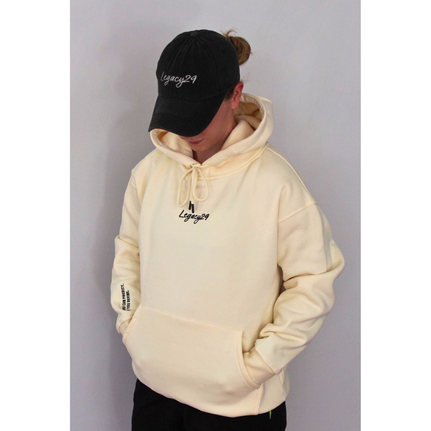 Person in Legacy29 Cloud 9 cream hoodie with hands in pockets, wearing a black cap. Cosy oversized fit with embroidered details for ultimate comfort.