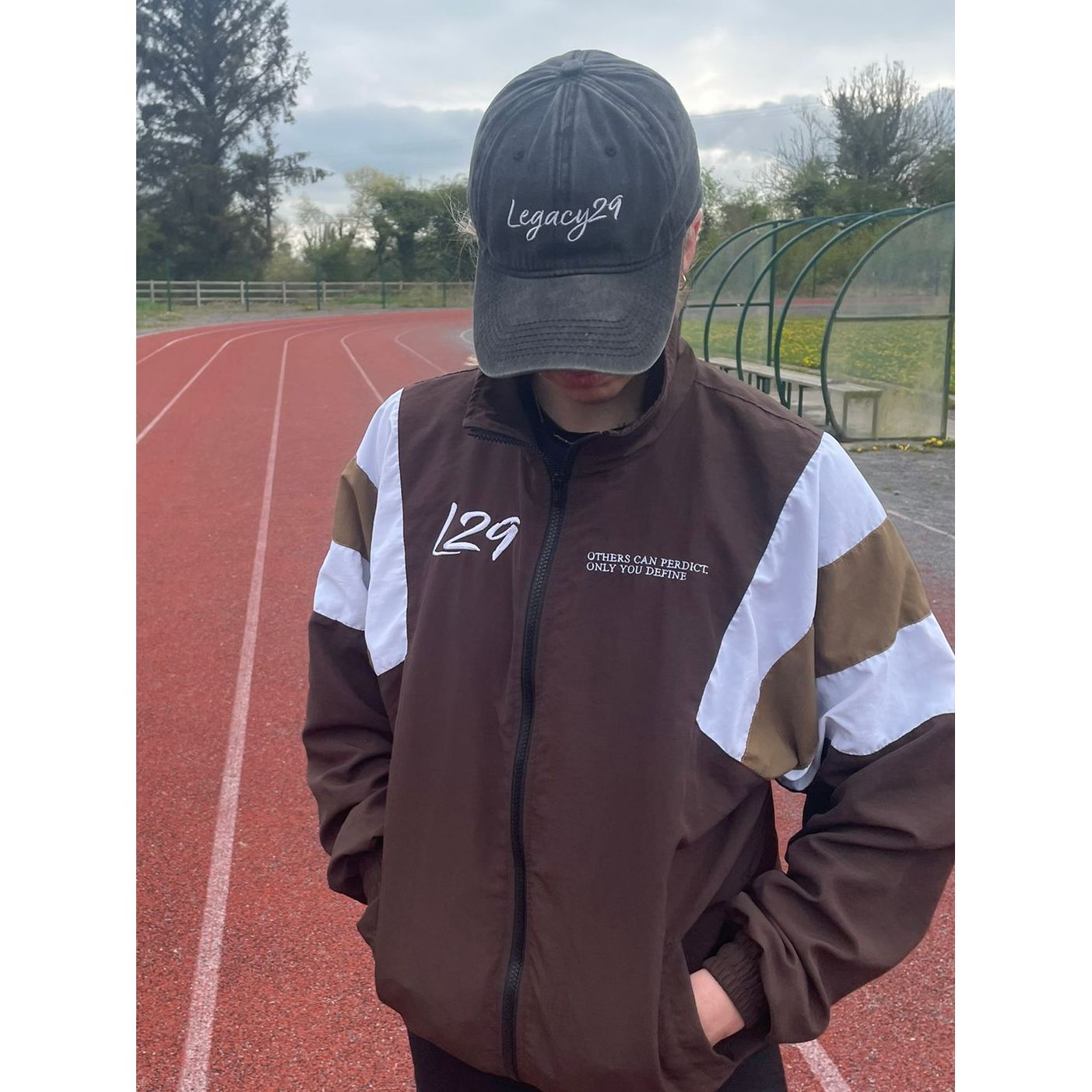 Person wearing a brown jacket with white accents and a black cap on a track field.